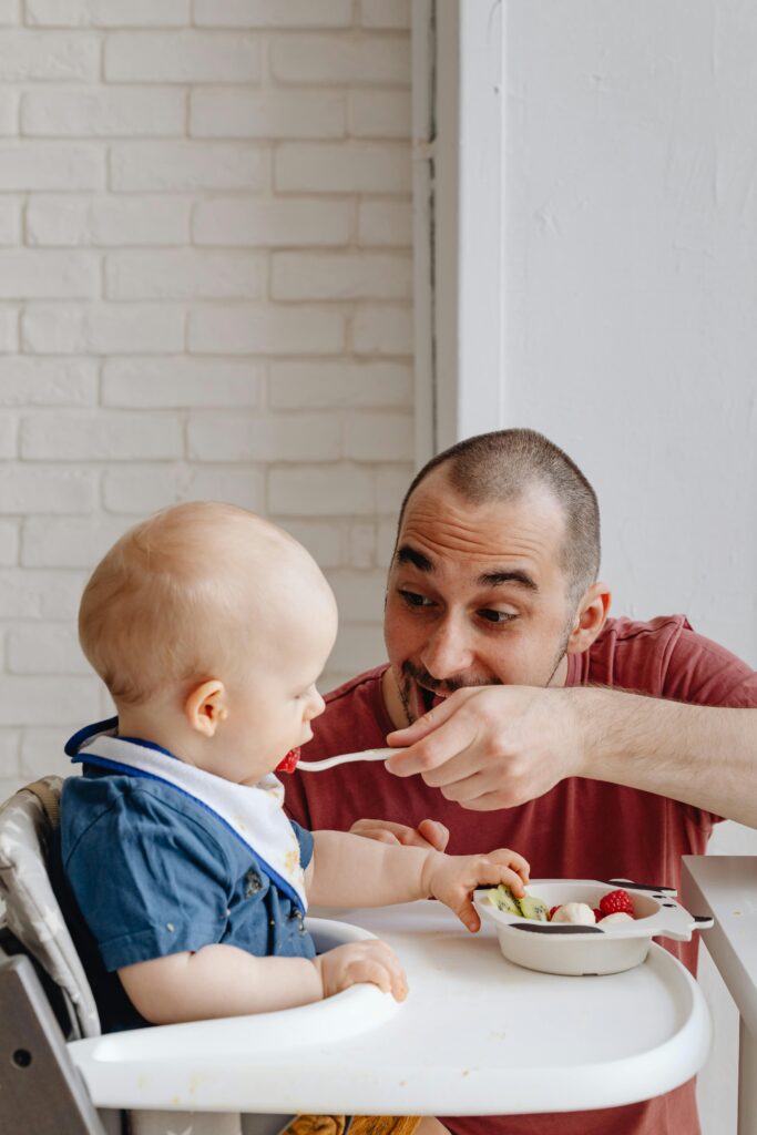 A father lovingly feeds his baby seated in a high chair against a brick wall backdrop indoors.